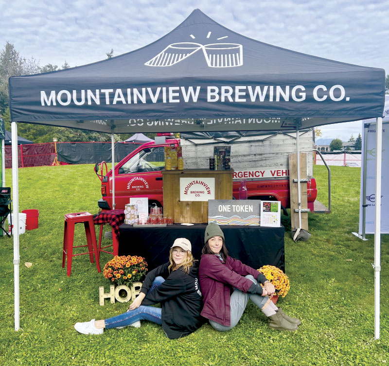 Two women sitting back-to-back in the grass in front of the Mountainview Brewing Co. booth