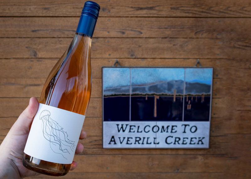Bottle of wine being held in front of Welcome to Averill Creek sign hanging on wall