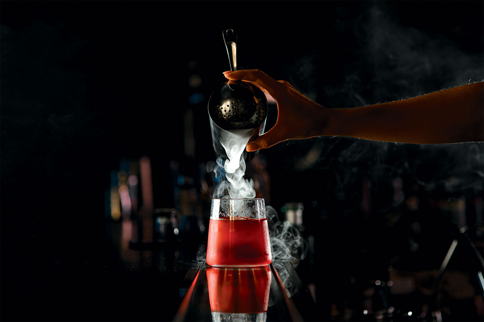 Bartender pours cocktail from steel shaker into drinking glass against dark blurred background